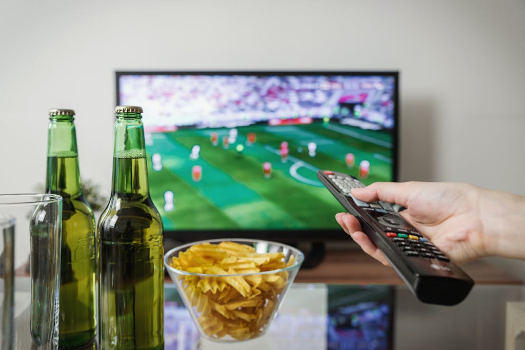 Serlig A person holding a remote while watching soccer on TV with beer and snacks on the table.