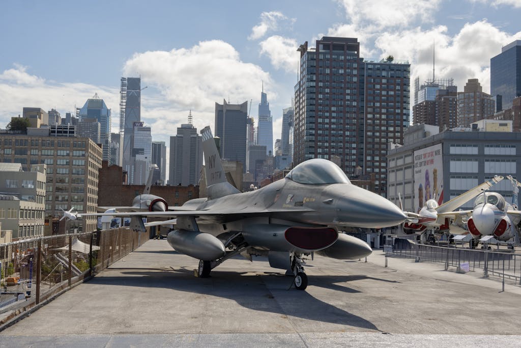 New York Jets F-16 fighter jet displayed at New York City's Intrepid Museum with skyline backdrop.