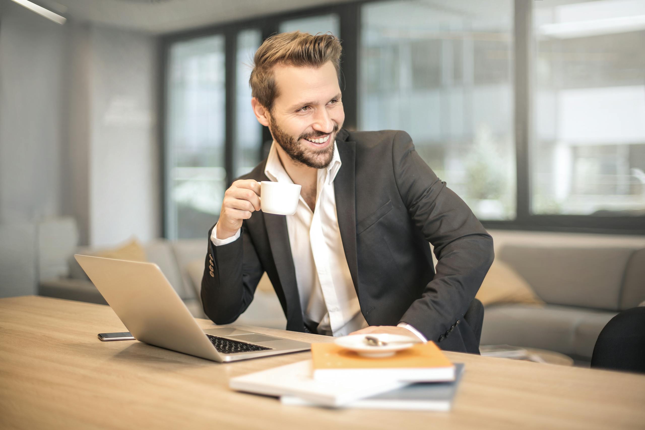 Entrepreneur Break Smiling businessman in formal attire enjoying coffee break at modern office desk with laptop.