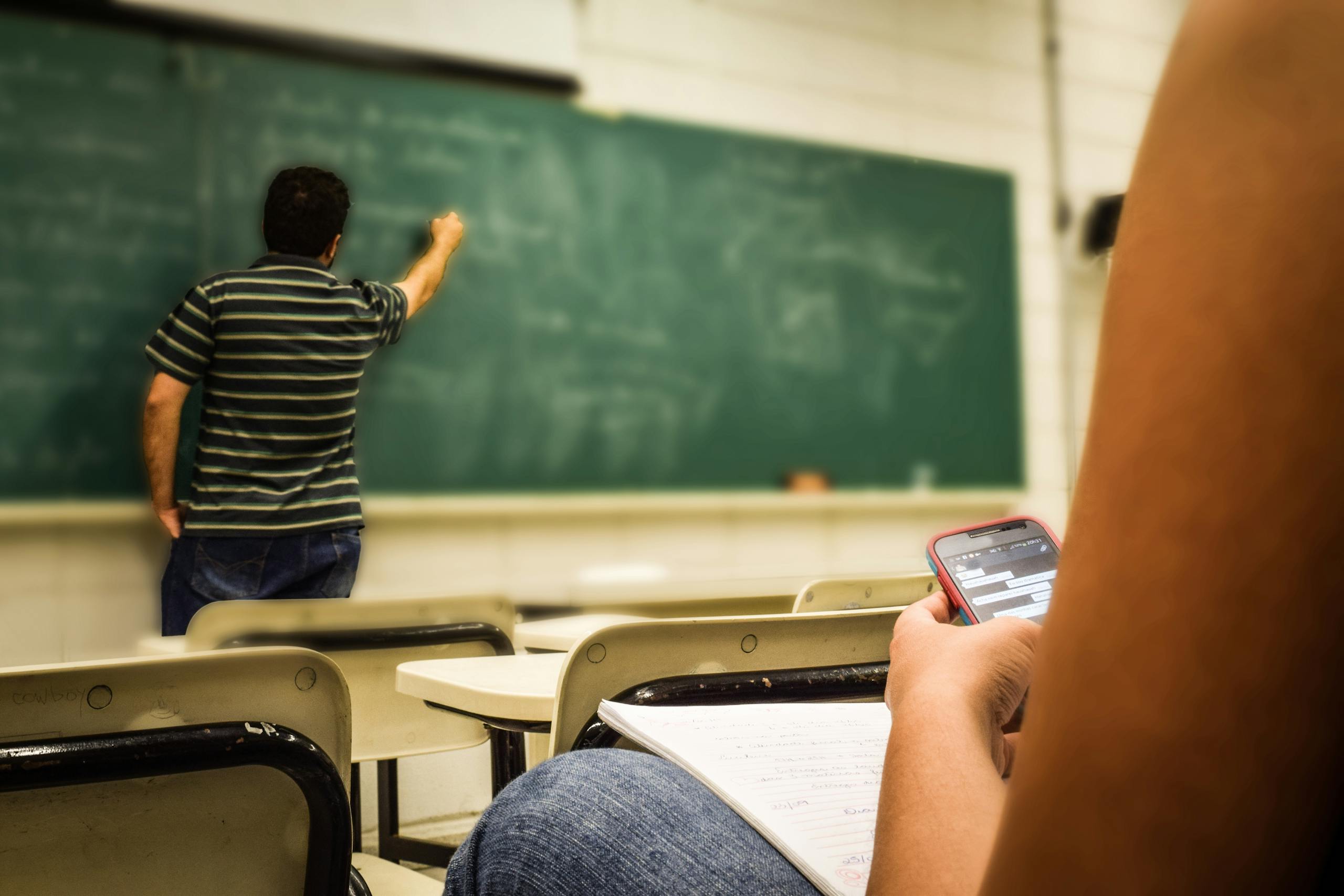 Algebra.learning Nexus.one Student texting in a classroom while teacher is writing on the blackboard.